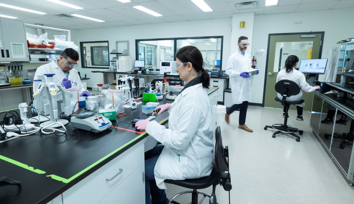 Scientists in white lab coats work around a black lab bench.