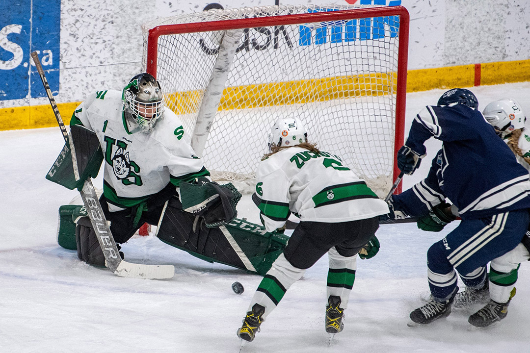 Goaltender Camryn Drever is seen in mid-game action, defending against the opposing hockey team at Merlis Belsher Place. Several other hockey players can be seen attempting to gain control of the puck in front of the net. 