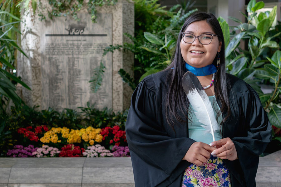 Portrait of Cree Autumn Sunrise Longjohn, wearing graduation garb and holding a feather. 
