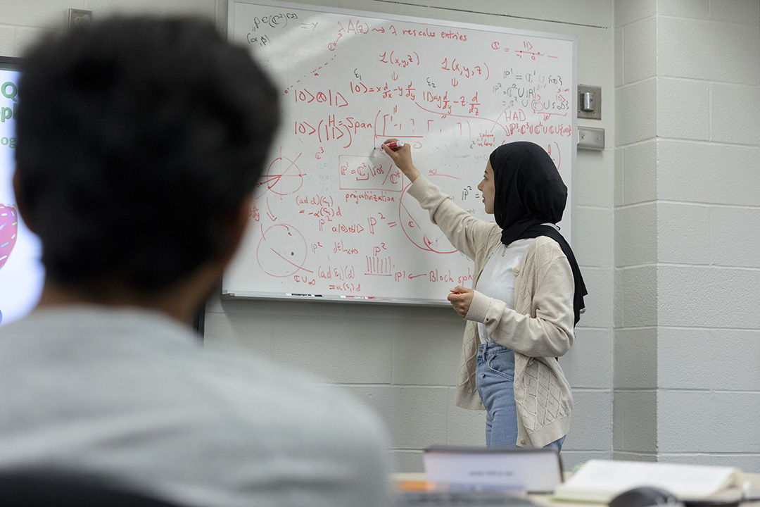 A student writes math equations on a white board in a classroom.