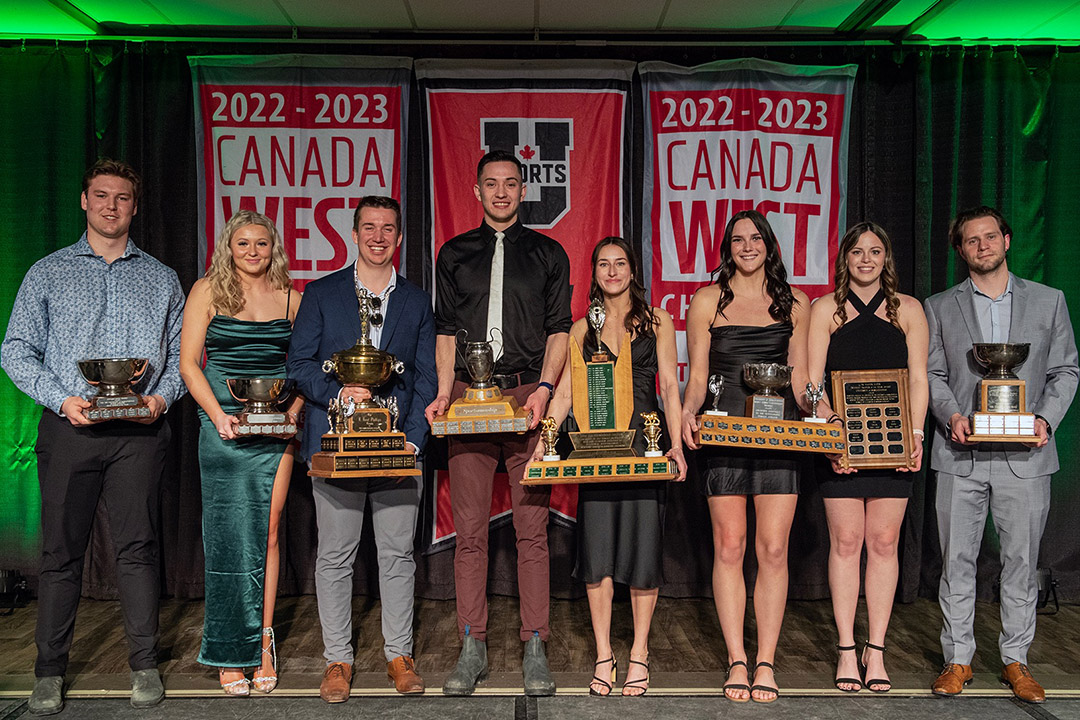 Eight student athletes holding awards at a ceremony with banners and green lights in the background.